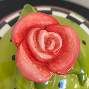 以下が含まれることがあります： 鮮やかな赤いバラと緑の葉が特徴のセラミック蓋のクローズアップ。 蓋は、黒い水玉模様の薄緑色のベースと、白と黒のチェック柄のボーダーが付いています。 バラが焦点です。