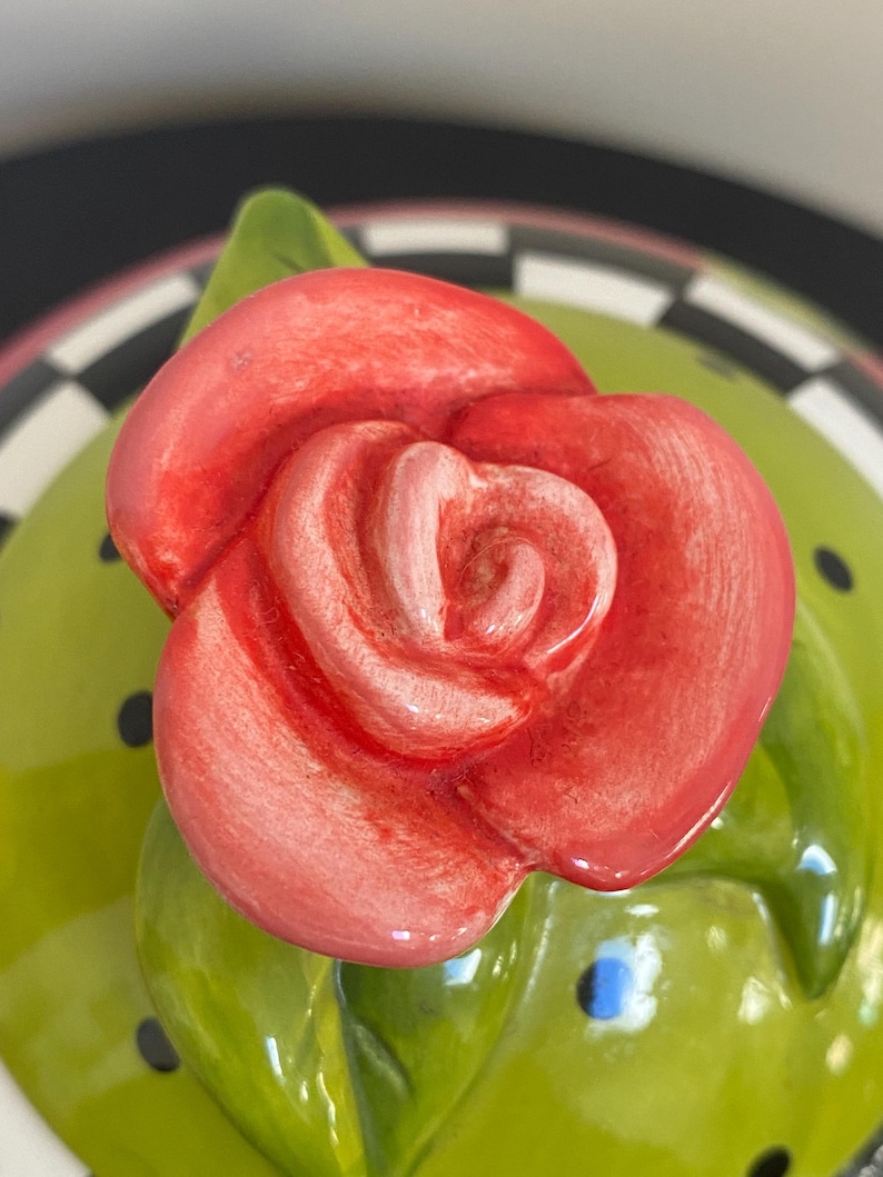 以下が含まれることがあります： 鮮やかな赤いバラと緑の葉が特徴のセラミック蓋のクローズアップ。 蓋は、黒い水玉模様の薄緑色のベースと、白と黒のチェック柄のボーダーが付いています。 バラが焦点です。