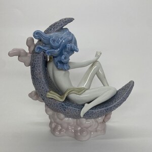 May include: A porcelain figurine of a seated figure with blue hair, resting on a crescent moon. The moon is a textured, lavender color, and the figure is draped with a cream-colored cloth. The base is a cloud-like formation in shades of pink.