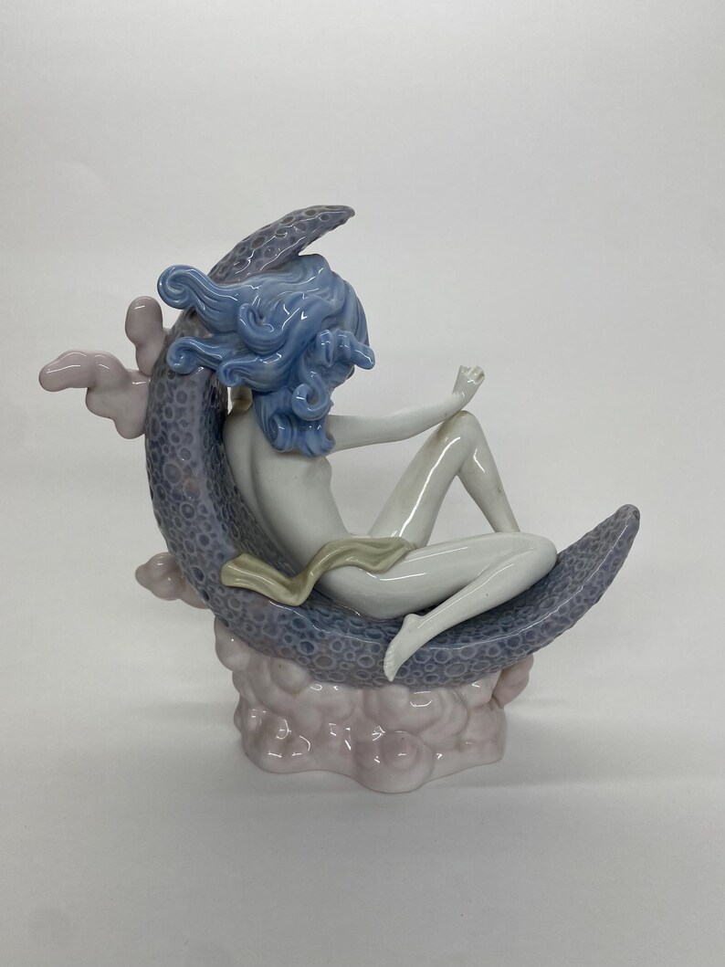 May include: A porcelain figurine of a seated figure with blue hair, resting on a crescent moon. The moon is a textured, lavender color, and the figure is draped with a cream-colored cloth. The base is a cloud-like formation in shades of pink.