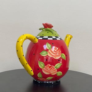 以下が含まれることがあります： 黒い水玉模様の黄色いハンドルが付いた、装飾的な赤いティーポット。 ティーポットにはバラのイラストと、緑と黒のチェック柄の蓋が付いています。赤いバラが蓋のハンドルとして機能します。