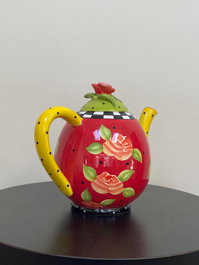 以下が含まれることがあります： 黒い水玉模様の黄色いハンドルが付いた、装飾的な赤いティーポット。 ティーポットにはバラのイラストと、緑と黒のチェック柄の蓋が付いています。赤いバラが蓋のハンドルとして機能します。