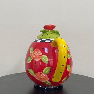 以下が含まれることがあります： 赤いセラミック製の装飾的なティーポット。黄色のハンドルと赤いバラの蓋が付いています。ティーポットは、描かれたバラと緑の葉、黒い水玉模様で飾られています。ベースは白と黒のチェック柄です。