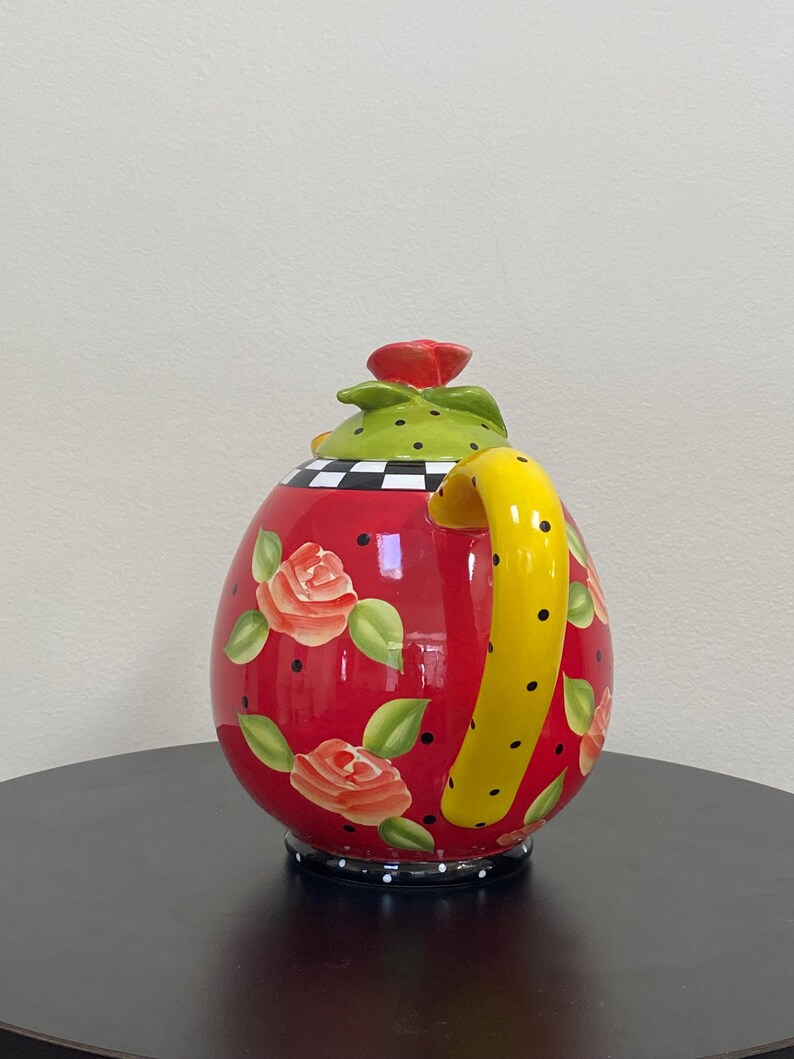 以下が含まれることがあります： 赤いセラミック製の装飾的なティーポット。黄色のハンドルと赤いバラの蓋が付いています。ティーポットは、描かれたバラと緑の葉、黒い水玉模様で飾られています。ベースは白と黒のチェック柄です。