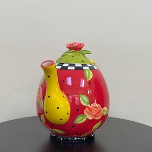 以下が含まれることがあります： 黄色い注ぎ口と取っ手が付いた、装飾的な赤いセラミックティーポット。 ティーポットは、上部に赤いバラ、黒い水玉模様、花のバラのアクセントが付いた緑色の蓋を備えています。 ベースには白と黒の市松模様があります。