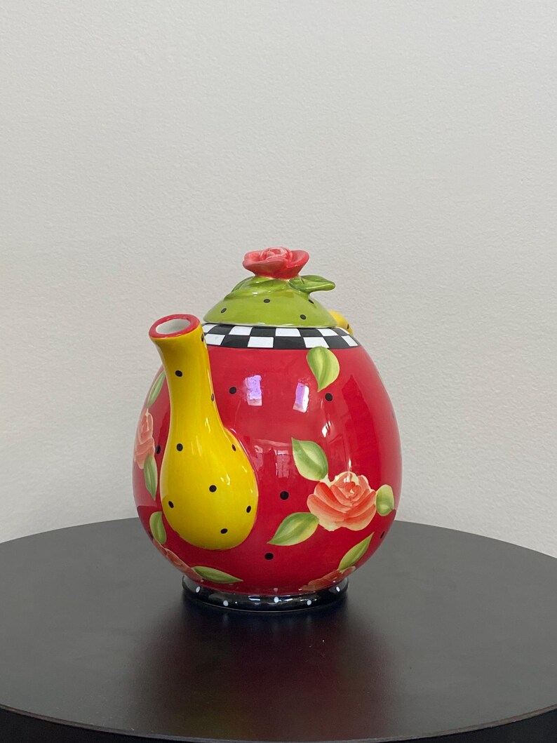 以下が含まれることがあります： 黄色い注ぎ口と取っ手が付いた、装飾的な赤いセラミックティーポット。 ティーポットは、上部に赤いバラ、黒い水玉模様、花のバラのアクセントが付いた緑色の蓋を備えています。 ベースには白と黒の市松模様があります。