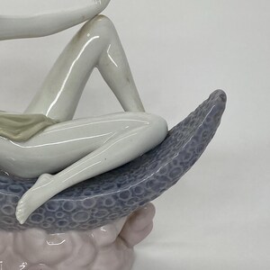 May include: A porcelain figurine depicting a reclining figure. The figure is primarily white with subtle shading, resting on a textured, blue-gray crescent shape. The base is a soft pink hue, creating a serene and artistic composition.