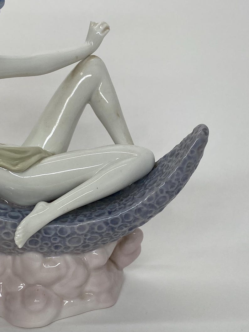 May include: A porcelain figurine depicting a reclining figure. The figure is primarily white with subtle shading, resting on a textured, blue-gray crescent shape. The base is a soft pink hue, creating a serene and artistic composition.