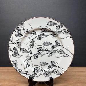 Peut inclure: Assiette en c&eacute;ramique blanche avec un motif floral noir et blanc. L'assiette a un bord dor&eacute; et est soutenue par un support noir. Le motif comprend des feuilles stylis&eacute;es et des motifs complexes, cr&eacute;ant une esth&eacute;tique &eacute;l&eacute;gante.