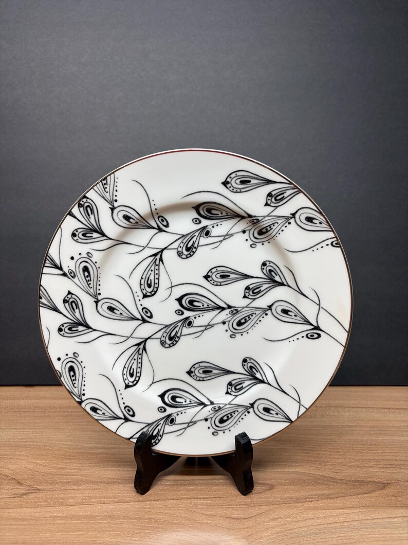 Peut inclure: Assiette en c&eacute;ramique blanche avec un motif floral noir et blanc. L'assiette a un bord dor&eacute; et est soutenue par un support noir. Le motif comprend des feuilles stylis&eacute;es et des motifs complexes, cr&eacute;ant une esth&eacute;tique &eacute;l&eacute;gante.