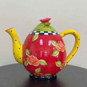 以下が含まれることがあります： バラのイラストと黒い水玉模様が施された赤いボディの装飾用セラミックティーポット。注ぎ口とハンドルは黒い水玉模様の黄色です。蓋は緑色で、赤いバラのつまみが付いています。ベースは白と黒の市松模様です。