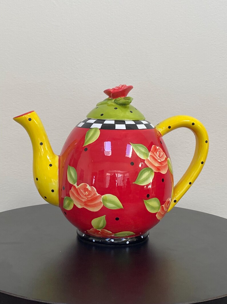 以下が含まれることがあります： バラのイラストと黒い水玉模様が施された赤いボディの装飾用セラミックティーポット。注ぎ口とハンドルは黒い水玉模様の黄色です。蓋は緑色で、赤いバラのつまみが付いています。ベースは白と黒の市松模様です。