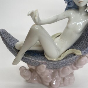 May include: A porcelain figurine of a reclining figure with blue hair, resting on a textured, blue and gray surface. The figure is nude, with a light green snake-like detail. The base is a light pink and gray cloud-like shape.