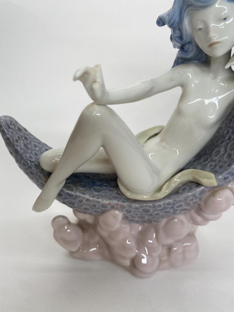 May include: A porcelain figurine of a reclining figure with blue hair, resting on a textured, blue and gray surface. The figure is nude, with a light green snake-like detail. The base is a light pink and gray cloud-like shape.