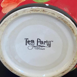 以下が含まれることがあります： 赤色の外側、黒い縁、白い内側を持つセラミックティーパーティーボウル。ボウルには手描きの赤いバラと緑の葉が描かれています。ボウルの底には「Tea Party」と「Joyce Sheen Giftcraft」の文字があります。