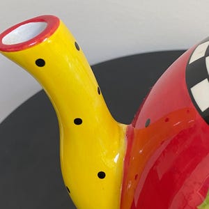 以下が含まれることがあります： 黄色い注ぎ口に黒い水玉模様が施された、カラフルなセラミックティーポット。 ティーポットの本体は赤色で、上部には白と黒の市松模様があります。 注ぎ口には赤い縁取りがあります。