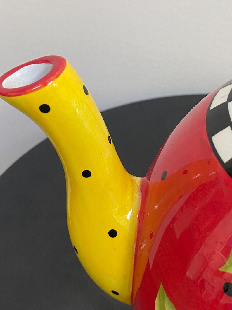 以下が含まれることがあります： 黄色い注ぎ口に黒い水玉模様が施された、カラフルなセラミックティーポット。 ティーポットの本体は赤色で、上部には白と黒の市松模様があります。 注ぎ口には赤い縁取りがあります。