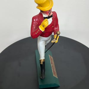 以下が含まれることがあります： 赤いジャケット、黄色の手袋、黄色のシルクハットを身に着け、杖を持っている男性のヴィンテージフィギュア。 フィギュアは「JOHNNIE WALKER」の文字が入った緑色の台座の上に立っています。 フィギュアの高さは約18cmです。