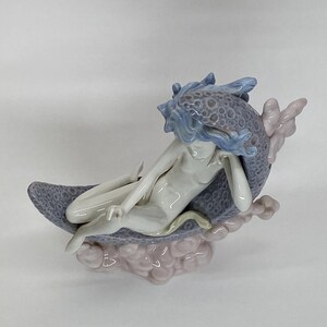 May include: A porcelain figurine of a nude figure reclining on a crescent moon. The figure has blue hair and is resting on a moon with a textured, speckled surface. The base is a cluster of pink clouds.