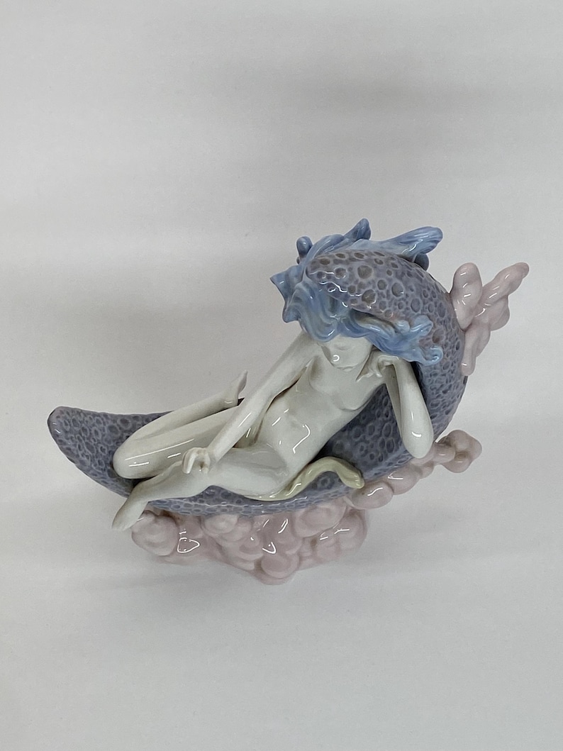 May include: A porcelain figurine of a nude figure reclining on a crescent moon. The figure has blue hair and is resting on a moon with a textured, speckled surface. The base is a cluster of pink clouds.