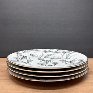 Peut inclure: Quatre assiettes en c&eacute;ramique blanche avec un motif de feuilles noir et blanc. Les assiettes ont un fin bord argent&eacute; et sont empil&eacute;es sur une surface en bois. Le fond est gris fonc&eacute;.