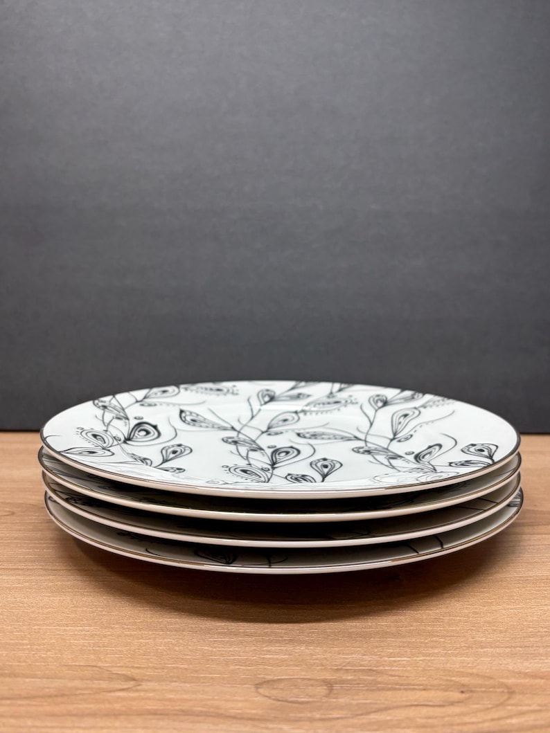 Peut inclure: Quatre assiettes en c&eacute;ramique blanche avec un motif de feuilles noir et blanc. Les assiettes ont un fin bord argent&eacute; et sont empil&eacute;es sur une surface en bois. Le fond est gris fonc&eacute;.