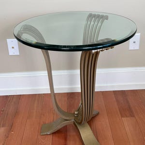 May include: A round glass-topped side table with a gold-colored, Art Deco-style base. The base features curved supports and vertical lines, and the table stands on a four-legged base. The table is approximately 24 inches in height.