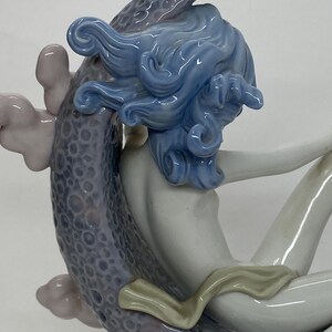 May include: A porcelain figurine of a woman with flowing blue hair, seated on a textured, lavender-colored form. The woman's skin is a pale, creamy white, and she wears a light-colored garment. The figurine has a glossy finish.