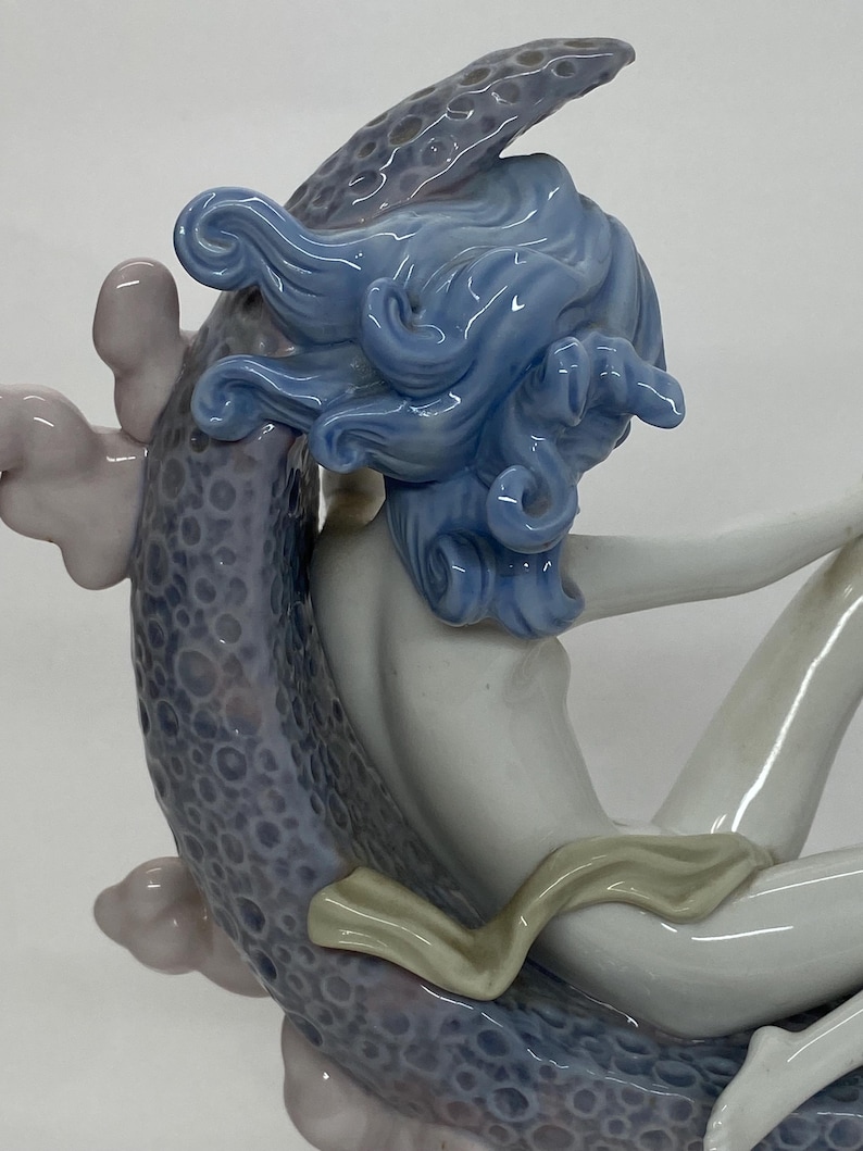 May include: A porcelain figurine of a woman with flowing blue hair, seated on a textured, lavender-colored form. The woman's skin is a pale, creamy white, and she wears a light-colored garment. The figurine has a glossy finish.
