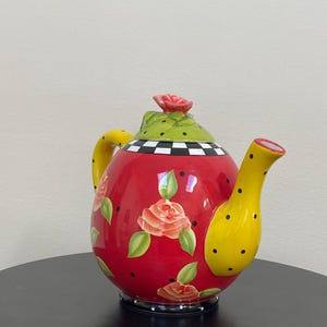 以下が含まれることがあります： 黄色い取っ手と注ぎ口が付いた、装飾的な赤いティーポット。 ティーポットは、オレンジ色のバラ、緑の葉、黒い水玉模様で飾られています。 蓋は黒い水玉模様と赤いバラが付いた緑色です。 ベースは白と黒の市松模様です。