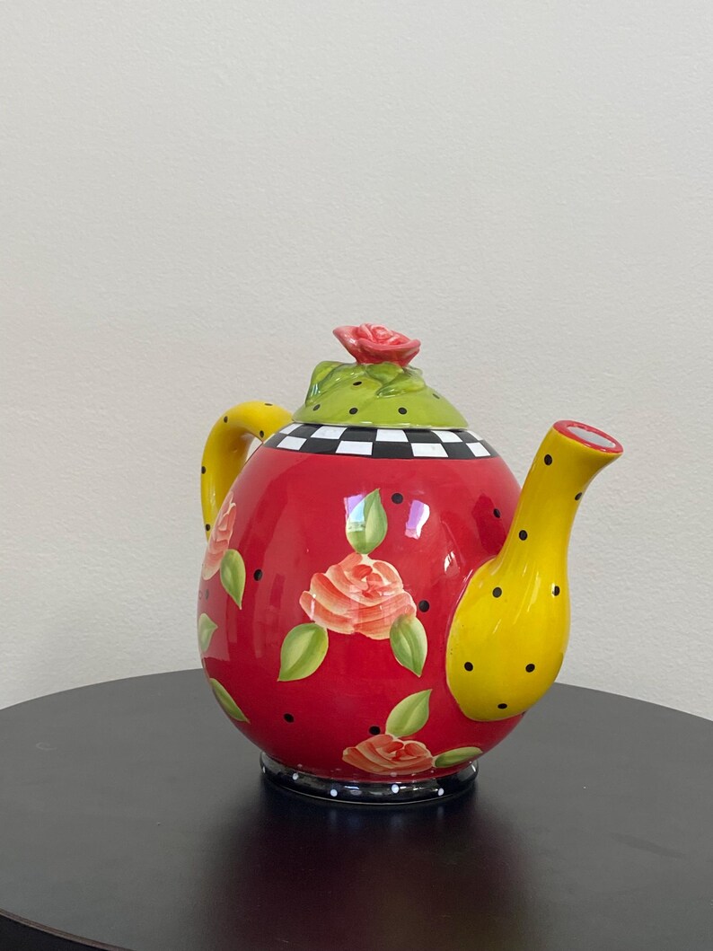 以下が含まれることがあります： 黄色い取っ手と注ぎ口が付いた、装飾的な赤いティーポット。 ティーポットは、オレンジ色のバラ、緑の葉、黒い水玉模様で飾られています。 蓋は黒い水玉模様と赤いバラが付いた緑色です。 ベースは白と黒の市松模様です。