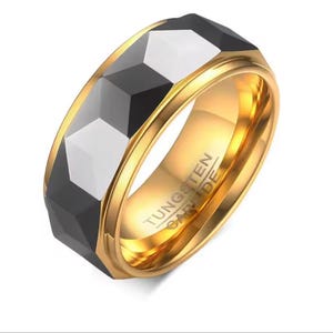 May include: A gold-coloured tungsten carbide ring featuring a faceted black and silver band. The ring has a polished gold interior, with the text "TUNGSTEN CARBIDE" visible on the inside of the band.