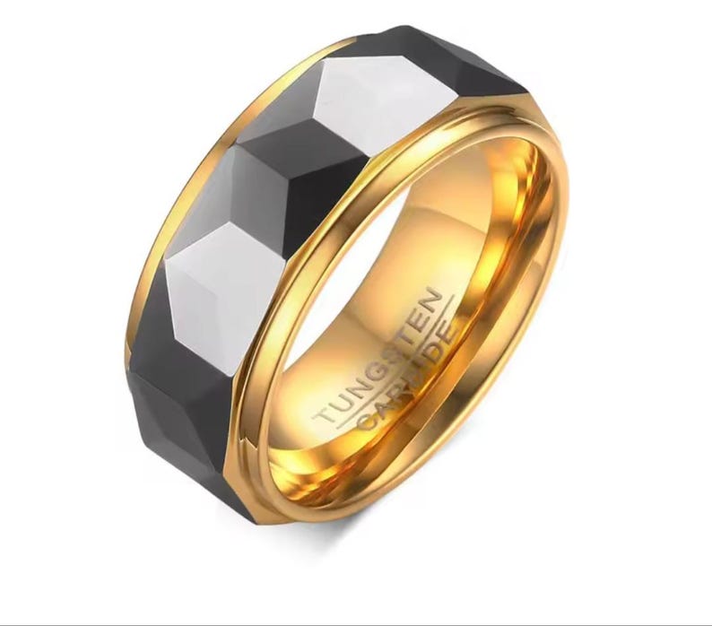 May include: A gold-coloured tungsten carbide ring featuring a faceted black and silver band. The ring has a polished gold interior, with the text "TUNGSTEN CARBIDE" visible on the inside of the band.