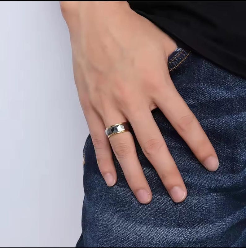May include: A gold-coloured ring with a geometric design and a silver and black centre band. The ring is worn on a finger, showcasing its polished finish and modern style. The ring's design features a combination of metallic colours.