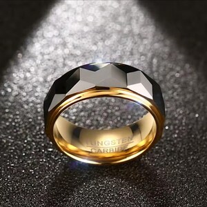May include: A gold-coloured tungsten carbide ring with a faceted black top. The ring has a polished gold interior and the words "TUNGSTEN CARBIDE" are engraved inside. The ring is displayed on a dark, textured surface.