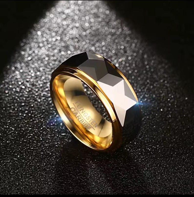 May include: A gold-coloured tungsten carbide ring with a faceted, geometric design. The ring has a polished finish and features the engraved text "Tungsten Carbide". The ring is displayed on a dark, textured surface.