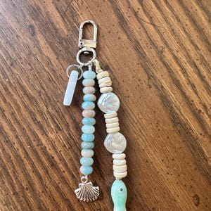 May include: A handmade keychain with a silver clasp and a variety of beads. The keychain features light blue and peach-colored beads, white shell-shaped beads, and a small, light green fish charm. A silver seashell charm hangs below the blue beads.