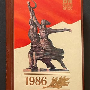 May include: A brown book with a red background and a statue illustration. The statue depicts a man and woman holding a hammer and sickle. The year 1986 is printed at the bottom. The text "XXVII" and "Съезд КПСС" are printed at the top.