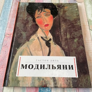 May include: A hardcover book featuring a portrait of a woman with red cheeks and lips, wearing a white shirt and black tie. The book's title, "MODIL'YANI," is displayed on a white rectangular label. The book has a red spine.