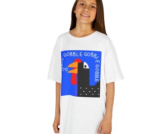 May include: White t-shirt featuring a cartoon turkey design with the words "GOBBLE GOBBLE" in blue. The turkey has a black body, red wattle, yellow beak, and white polka dots. The shirt is worn by a person.