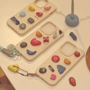 May include: Beige phone cases adorned with colorful, textured climbing holds. Each case features a unique arrangement of miniature holds in various shapes and colors, including yellow, red, blue, and gray. The cases are designed to protect the phone while adding a unique aesthetic.