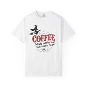 Puede incluir: Una camiseta blanca de manga corta con un gráfico de una bruja en una escoba sobre el texto rojo COFFEE y el texto negro helping witches and bitches since 1692, rodeado de pequeñas estrellas, lunas y una ilustración de una taza de café.