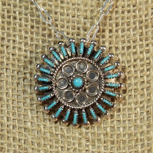 May include: A silver-toned pendant necklace with a circular design. The pendant features a central turquoise stone surrounded by smaller turquoise accents and silver beads. The necklace hangs on a silver chain.