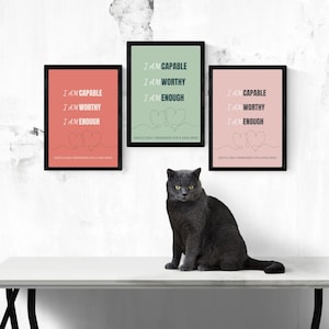 Puede incluir: Tres impresiones enmarcadas con el texto "I AM CAPABLE, I AM WORTHY, I AM ENOUGH" en negro. Las impresiones son en coral, verde menta y rosa rubor. Un gato gris está sentado en una mesa blanca frente a las impresiones.