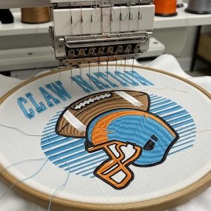 May include: An embroidery machine in operation, stitching a design onto white fabric. The design features a football and a helmet in shades of brown, blue, and orange, with the text "CLAW NATION" in blue. The fabric is stretched within a wooden hoop.