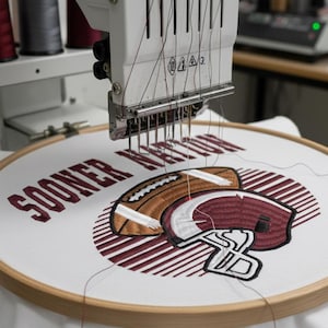 May include: An embroidery machine stitching a design onto white fabric stretched in a wooden hoop. The design features a football and helmet with the words "SOONER NATION" in maroon thread. Spools of thread in various colors are visible.
