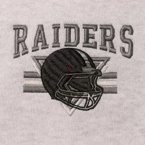 Puede incluir: Diseño bordado sobre tela gris claro. El diseño presenta la palabra "RAIDERS" en gris, arqueada sobre un casco de fútbol americano negro con detalles grises y blancos. El casco está posicionado sobre un triángulo gris y blanco.