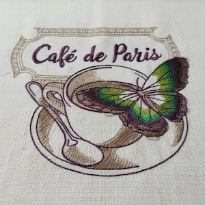 Peut inclure: Broderie représentant une tasse de café avec un papillon et le texte "Café de Paris". Le papillon a des ailes vertes et violettes. Le motif est dans les tons de marron et de violet sur fond blanc.