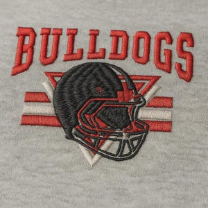 May include: Embroidered design featuring the word "BULLDOGS" in red, above a black and red football helmet. The helmet is set against a backdrop of red and white stripes and a white triangle. The design is on a light gray fabric.