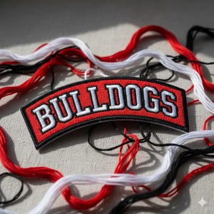 Pode incluir: Um patch bordado vermelho, preto e branco com a palavra "BULLDOGS" em letras brancas. Fios vermelhos, brancos e pretos circundam o patch, sugerindo um tema de artesanato ou costura.